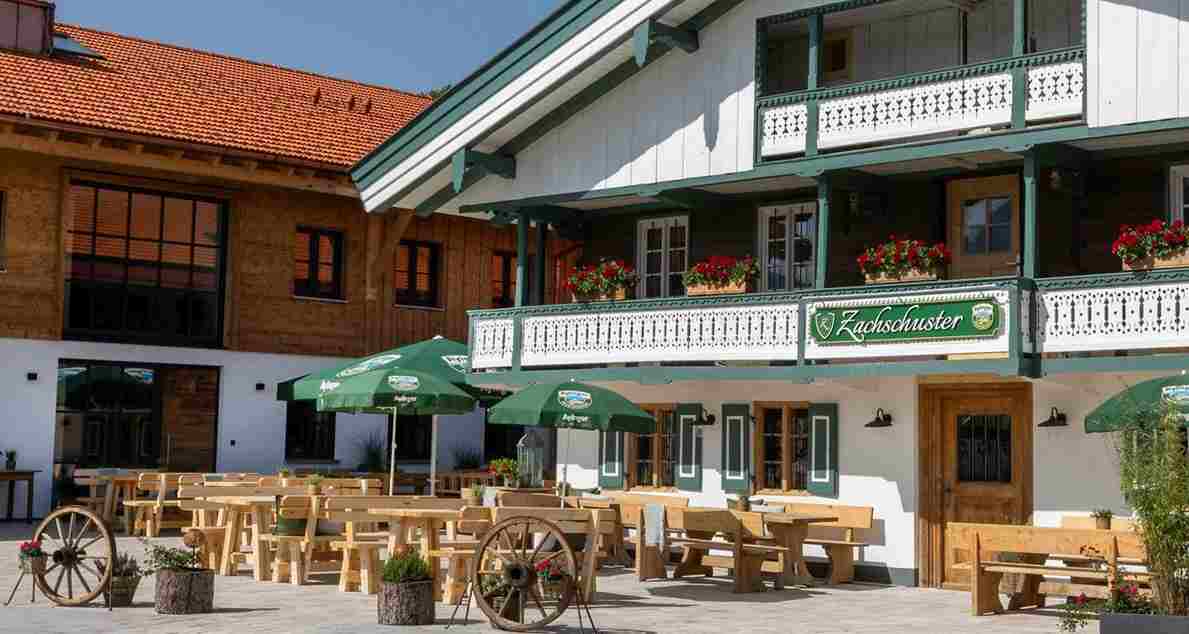 Sonnige Außenansicht des Landgasthofs Zachschuster in Gaißach mit weiß-grünem bayerischen Bauernhaus, Blumenkästen mit roten Geranien, Holzterrasse mit Biergarten-Bänken, grünen Sonnenschirmen und dekorativen Holz-Wagenrädern – vorbereitet als gemütliche Hochzeits-Location im Freien.