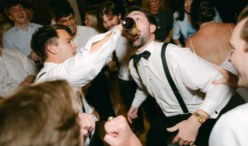 Ausgelassene Hochzeitsparty im Landgasthof Zachschuster, bei der mehrere Gäste in Hemd und Hosenträgern einen lachenden Bräutigam umringen, während ihm ein Freund aus einer Bierflasche direkt in den Mund einschenkt – dynamische Tanzfläche, pure Feierlaune und mitreißende Stimmung.