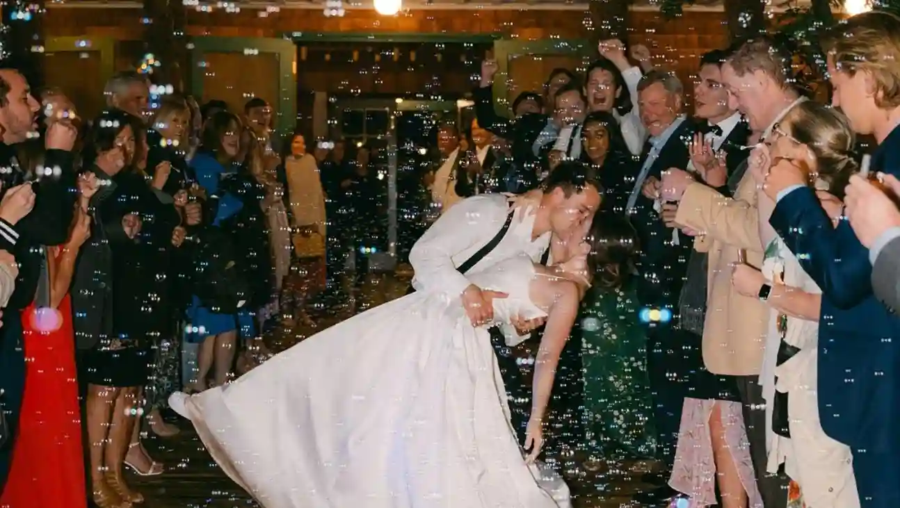 Romantischer Hochzeitsauszug bei Nacht in Gaißach: Das Brautpaar küsst sich in einem tiefen Dip, während es über einen Holzsteg zwischen zwei Reihen jubelnder Gäste läuft, die Seifenblasen in die Luft pusten und so einen funkelnden Tunnel aus Licht und Blasen bilden.