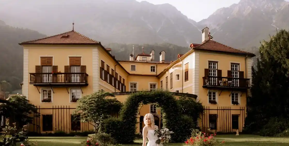Außenansicht des barocken Schloss Mühlau bei Innsbruck mit Satteldach, Fresken und weitläufigem Park unter blauem Himmel.