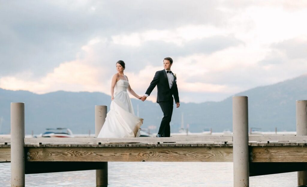 Braut und Bräutigam gehen Hand in Hand bei der Hochzeit über einen Holzsteg an einem See in Oberbayern, im Hintergrund Berge und ein dramatischer Himmel bei Sonnenuntergang.