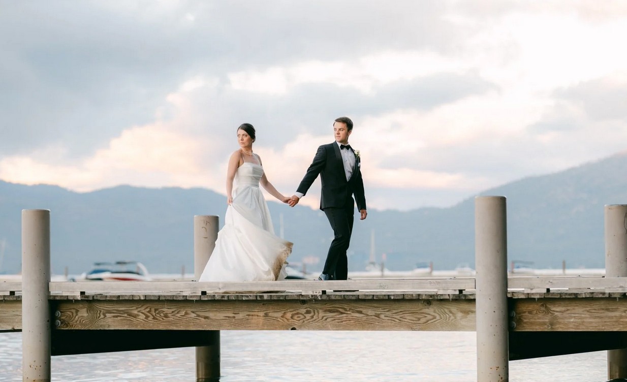 Braut und Bräutigam gehen Hand in Hand bei der Hochzeit über einen Holzsteg an einem See in Oberbayern, im Hintergrund Berge und ein dramatischer Himmel bei Sonnenuntergang.